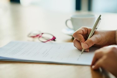 A male hand signing a document on a table next to spectacles and a cup of coffee.